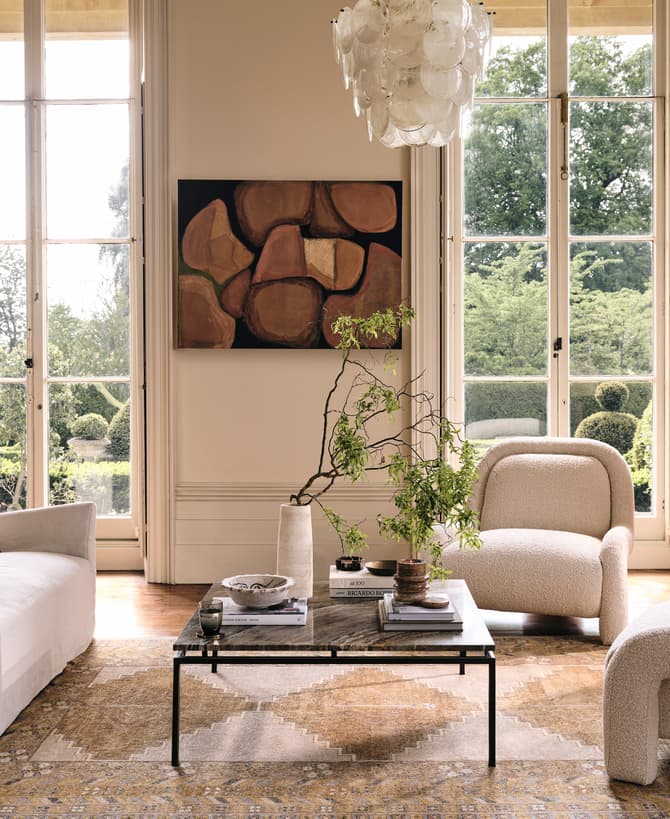 A living room with a white sofa, two armchairs, a coffee table with plants and books, surrounded by large windows overlooking a green garden, and decorated with abstract art and a chandelier.