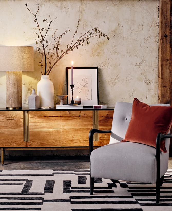 A contemporary living room features a grey armchair with a brown pillow, wooden sideboard with decor, including a lamp, plant, and candles, against a textured wall.