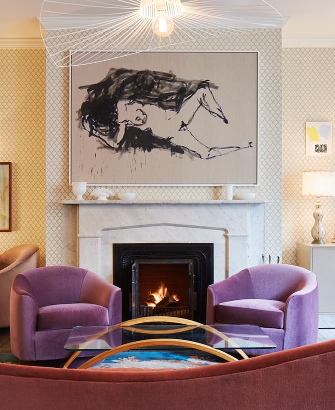 Cozy living room featuring colorful armchairs arranged around a glass coffee table, a lit fireplace with a large abstract painting above it, and art-adorned walls with warm lighting.