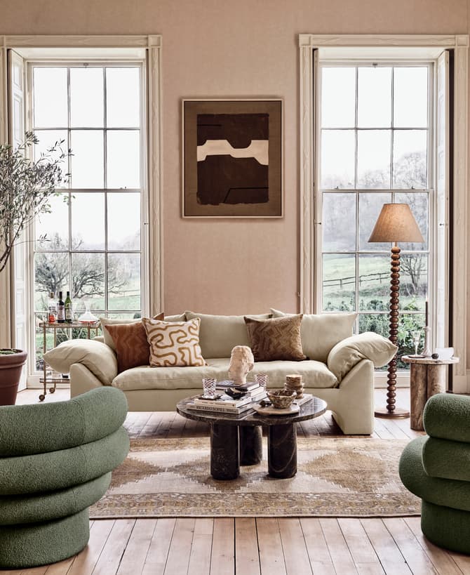 Modern living room with green, cylindrical chairs and beige sofa arranged around a central coffee table, accented by a potted tree, floor lamp, artwork, and large windows overlooking a pastoral landscape.