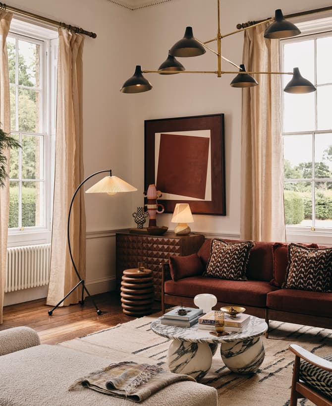 A cozy living room features a red sofa with patterned cushions, modern floor and ceiling lamps, a marble coffee table, a potted plant, and large windows with beige curtains.