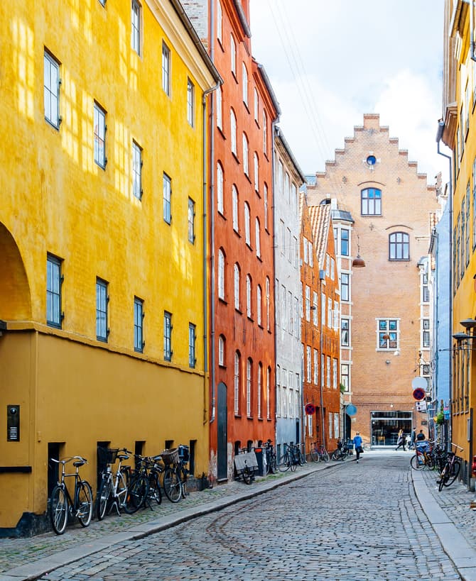 Bicycles line buildings with colorful facades—yellow, orange, blue, and brick—along a narrow cobblestone street in a quaint European city, under a bright, clear sky.