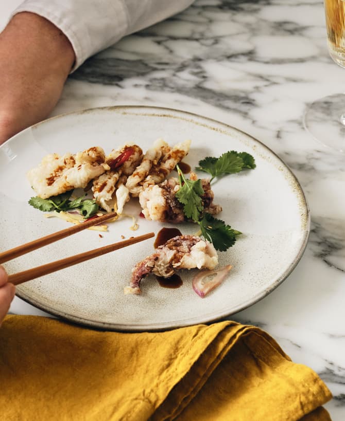 A person uses chopsticks to pick up a piece of fried food on a white plate garnished with greens, set on a marble table with a yellow napkin and a glass of white wine.