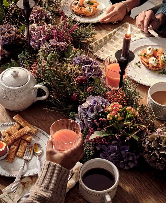 Table set for brunch with tea, juice, soft-boiled eggs, bread, and greenery centerpiece. People using utensils eating in a cozy, warmly lit environment. Plates and cups neatly arranged.