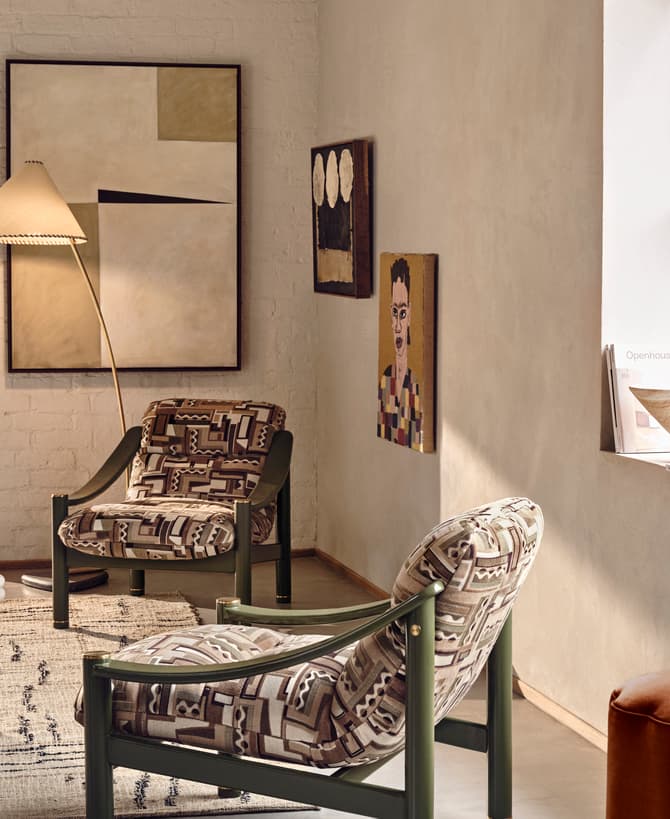 Two patterned armchairs sit in a cozy living room with modern art on the walls. A large window overlooks greenery, and a marble coffee table holds books and a glass.