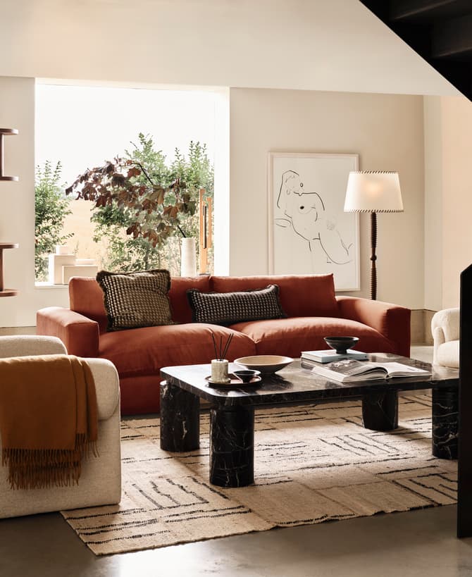 A modern living room with a red sofa, white armchair, and black marble coffee table; a bookshelf with decor, a framed sketch, and staircase in the corner; green plants seen outside.