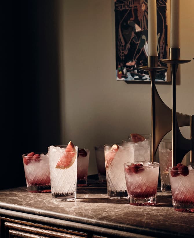 Several glasses filled with ice, raspberries, and a pink liquid rest on a marble-topped table. Behind them, a brass candelabra with white candles and a framed artwork adorn the wall.