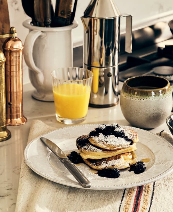 Pancakes topped with powdered sugar and berries sit on a white plate with a knife beside it, surrounded by a cup of coffee, a glass of orange juice, milk bottle, and cooking utensils in a kitchen.