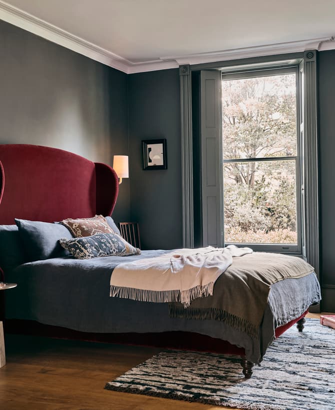 Luxurious bed with a red velvet headboard, adorned with neatly arranged pillows and a blanket, situated in an elegant, dark-walled bedroom featuring large windows with scenic views and modern décor.