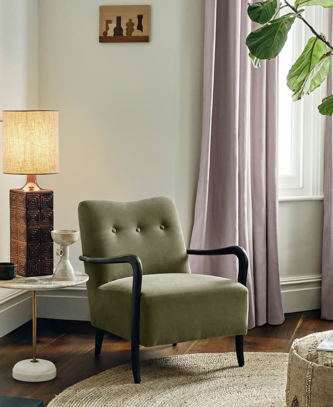 An olive-green armchair sits by a round side table with a lit lamp and decor items, placed next to a fireplace and in front of large windows with lavender curtains. There is a woven pouf with a magazine on top and a large indoor potted plant nearby.