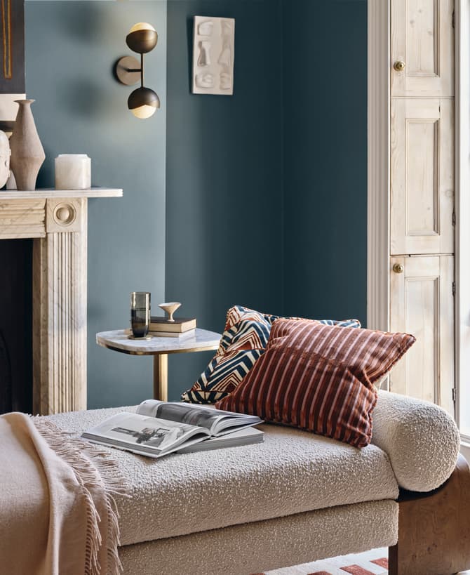 A chaise lounge with pillows and a blanket sits beside an open book and table with a glass, within a cozy room featuring a fireplace, wall art, and an open window with greenery outside.