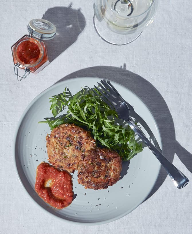 Two vegetable patties rest on a plate with arugula and red sauce, accompanied by a jar of red jam and a glass of water, on a white tablecloth. A fork is on the plate.
