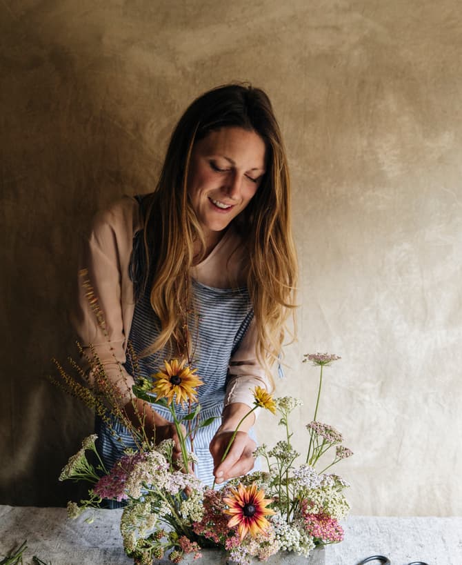 A person arranges a bouquet of flowers in a cement vase on a table, wearing an apron, with scissors nearby, against a textured beige wall.