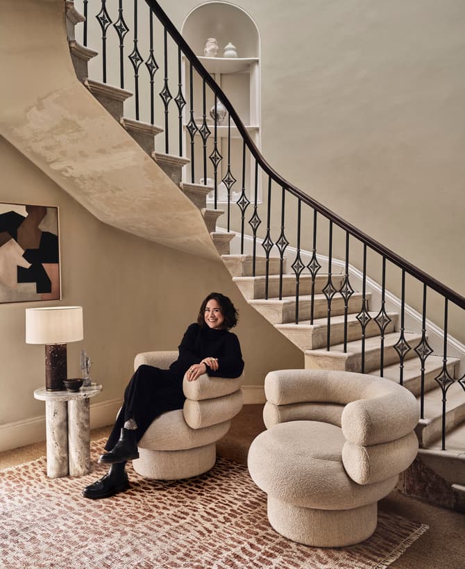 A person in black clothing sits smiling on a cushioned armchair beside a lamp table, under a staircase with an ornate railing. The room features a rug and a minimalist decor.