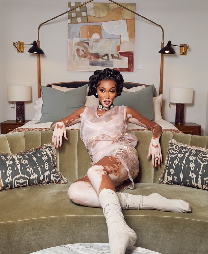 A woman lounges on a green velvet sofa in a softly lit, modern room with abstract art, wall-mounted lamps, and a marble coffee table. She's wearing a sheer, pink dress and cozy socks.