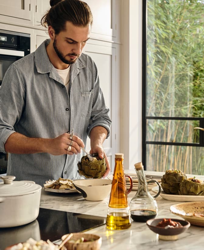 A person prepares artichokes at a kitchen counter, surrounded by bowls and bottles of oil and vinegar, near a window with a view of greenery.