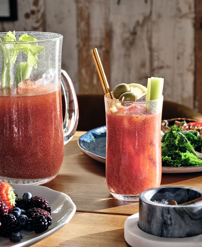 A pitcher and glass of red beverage, garnished with celery and olives, are on a wooden table surrounded by a salad, berries, and a bowl of salt.