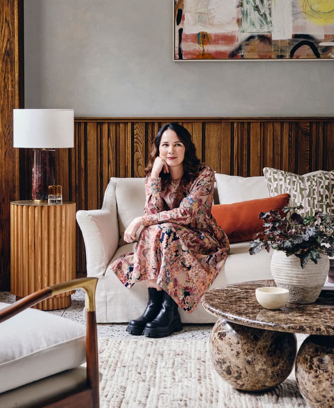 A woman in a floral dress sits on a beige sofa, resting her chin on her hand in a stylish living room with wooden paneling, a modern lamp, and abstract artwork above.