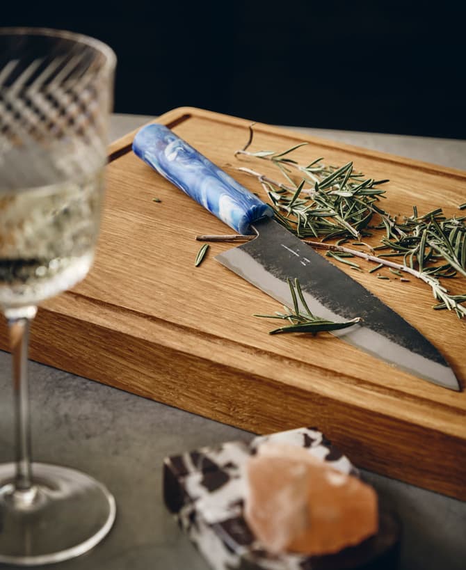 A knife with a blue handle lies on a wooden cutting board surrounded by fresh rosemary, next to a glass of white wine and a salt rock on a marble coaster.