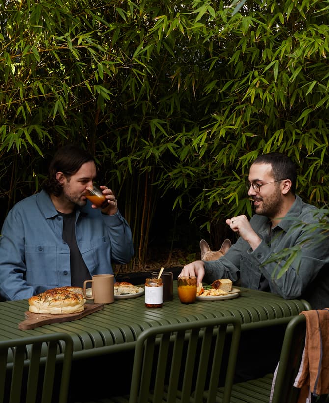 Two people sit at a green outdoor table eating and drinking, surrounded by dense bamboo foliage. A loaf of bread and jar with a white label are on the table.