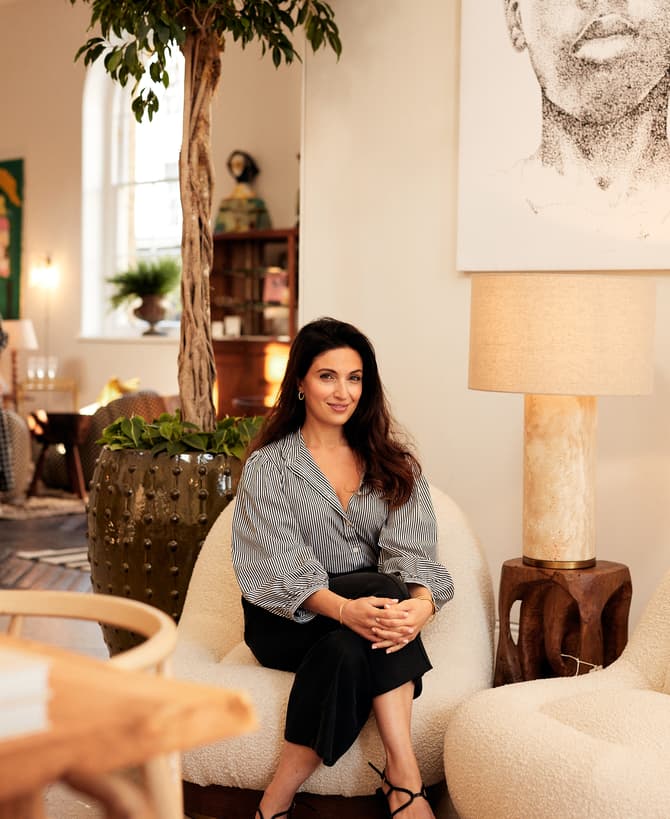 A woman with long dark hair and a striped blouse sits on a white chair in a well-decorated room with plants, modern furniture, and large windows; a large portrait is displayed on the wall.