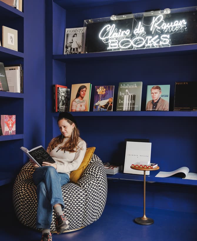 A woman reads a book while sitting in a patterned chair within a dark blue bookstore. The backdrop includes shelves with various books and a neon sign that reads "Claire de Rouen BOOKS."