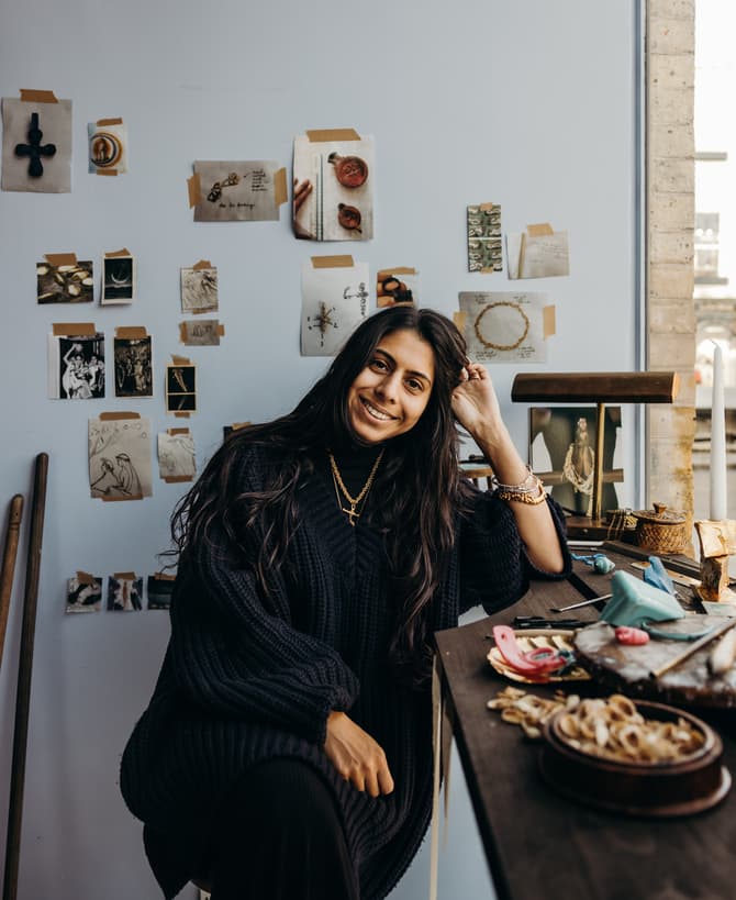 A woman with long hair and a blue sweater sits at a desk, smiling, with artwork and photos on the wall behind her in a well-lit room by a window.