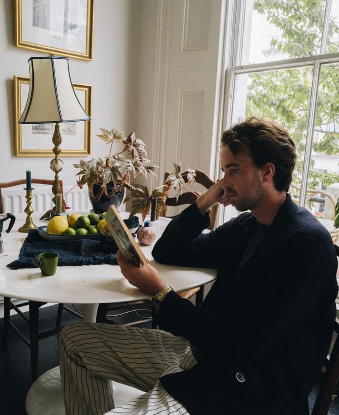 A man seated at a round white table reads a book; the table holds a lamp, plant, and a bowl of fruit beneath framed artworks and beside a large window.