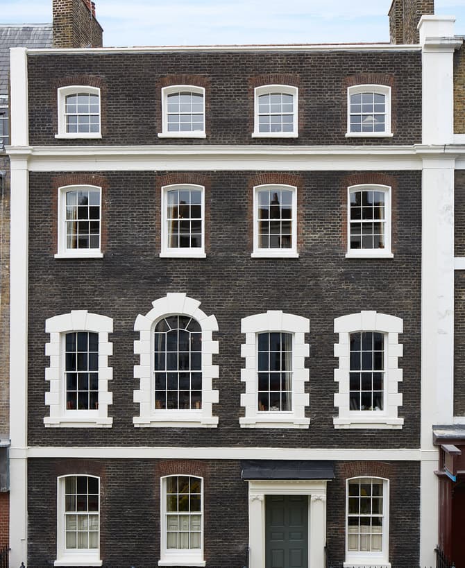 A four-story building with dark brickwork and white window frames stands in an urban setting. It features multiple symmetric windows and a green door on the ground floor.