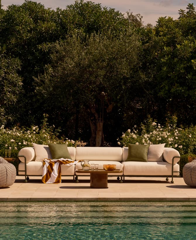 A white couch with green pillows and a striped blanket rests by a poolside, with a wooden table nearby holding glasses and a bowl; surrounded by lush green bushes and trees.