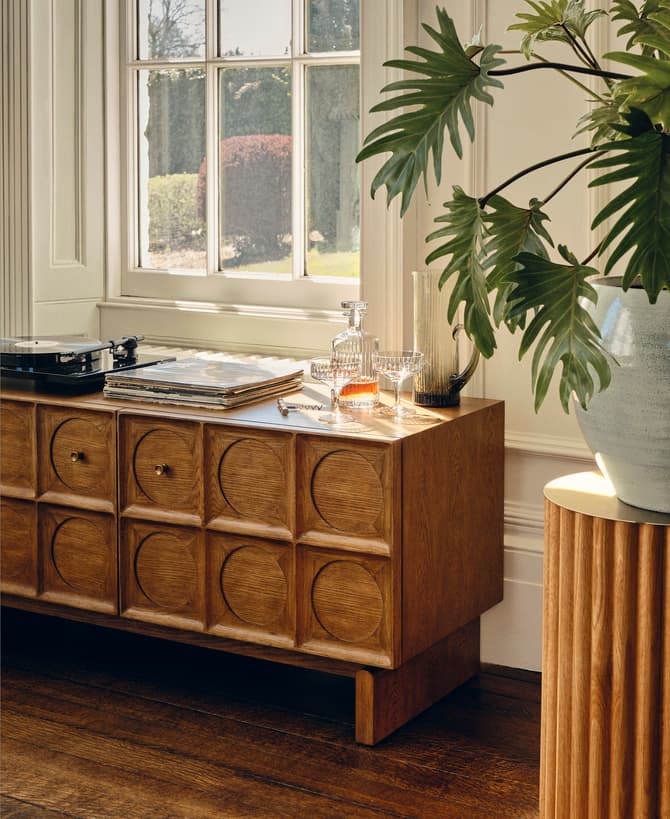 A wooden sideboard with geometric circular patterns holds vinyl records, a turntable, and glass decanters. It sits beside a window overlooking a garden, with a large potted plant nearby.