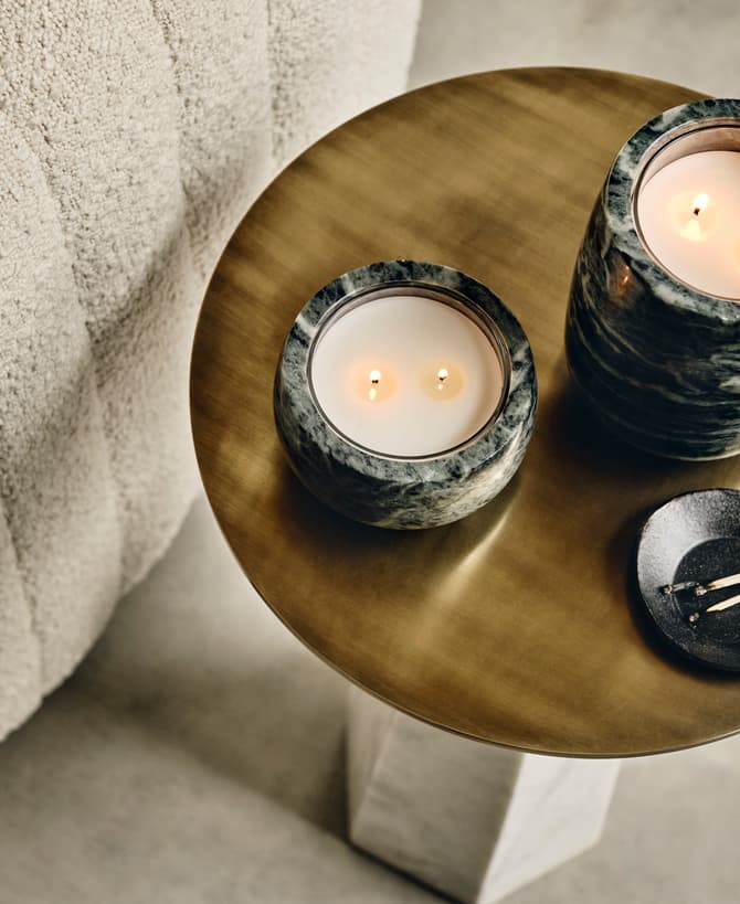 Candles are burning on a marble tabletop beside a textured, beige fabric sofa; a small black dish with matchsticks sits nearby.