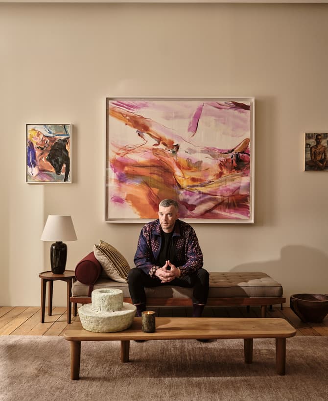A man sits on a bench in a living room, surrounded by abstract art on beige walls, with a wooden coffee table and decor items in the foreground.
