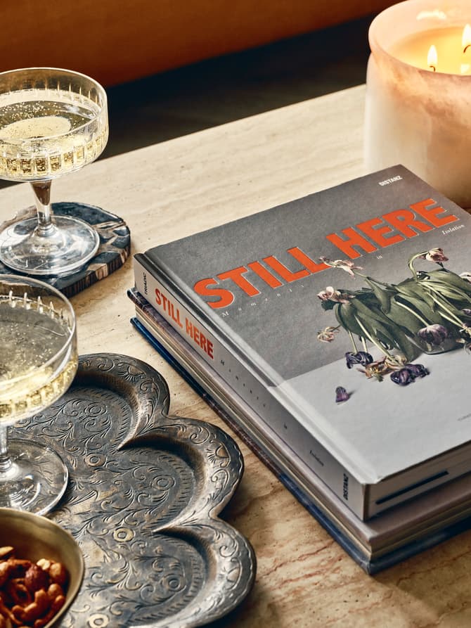 Two champagne flutes on coasters, a lit candle, a brass bowl of nuts, and books titled "STILL HERE" rest on a carved metal tray and a wooden table.