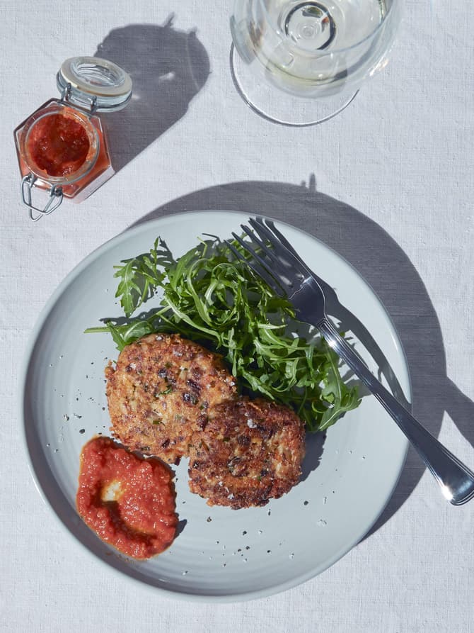 Two vegetable patties rest on a plate with arugula and red sauce, accompanied by a jar of red jam and a glass of water, on a white tablecloth. A fork is on the plate.