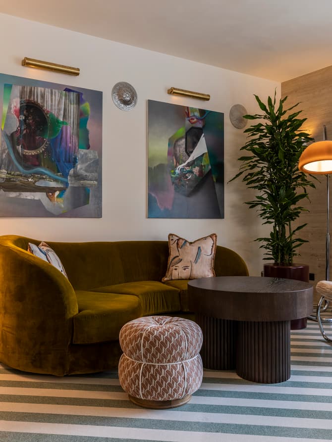 Mid-century modern living room featuring a green velvet sofa with decorative pillows, abstract wall art, a geometric table lamp, a striped ottoman, a round coffee table, a potted plant, and a unique lounge chair.