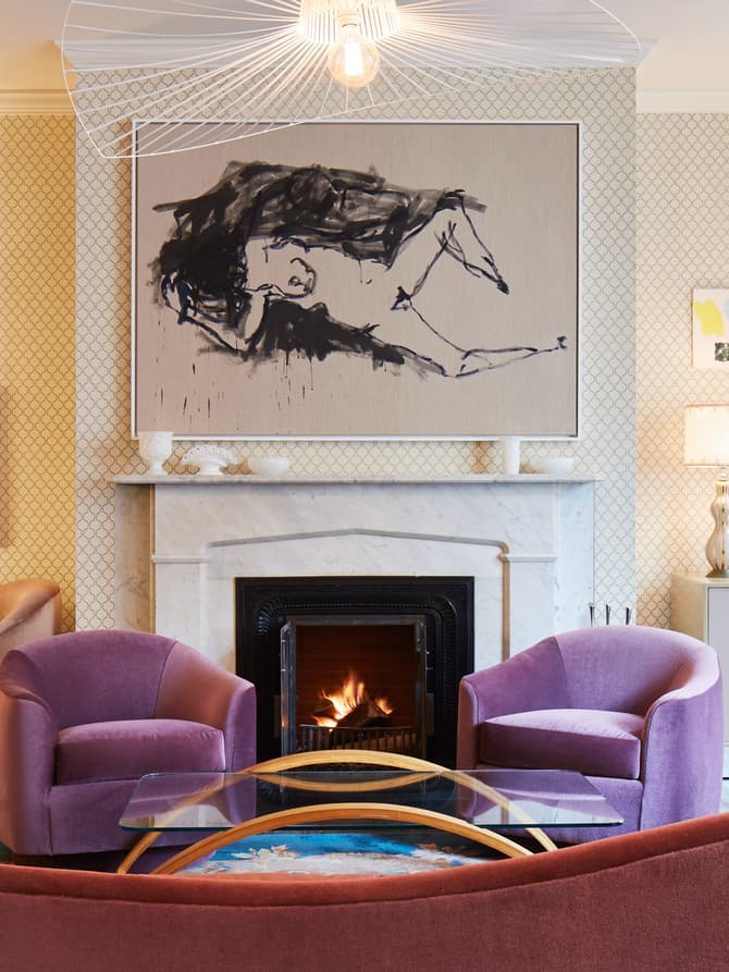 Cozy living room featuring colorful armchairs arranged around a glass coffee table, a lit fireplace with a large abstract painting above it, and art-adorned walls with warm lighting.