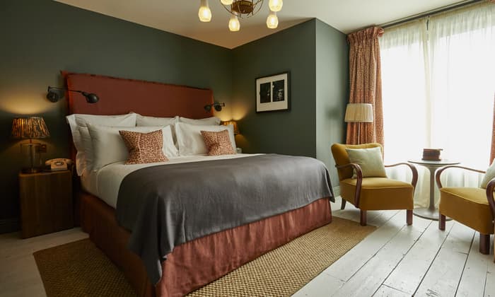 A cozy bedroom with a large bed, adorned with white linens and patterned pillows, sits under a chandelier; accompanied by a side table, armchairs, and a window with curtains.