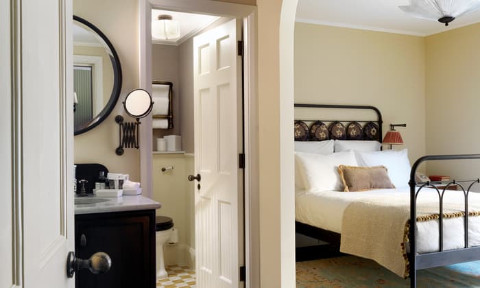 A neatly arranged bedroom with an ornate bed frame, a center pillow, and a cozy blanket is adjacent to an open bathroom door showing a vanity sink, round mirror, and toilet.