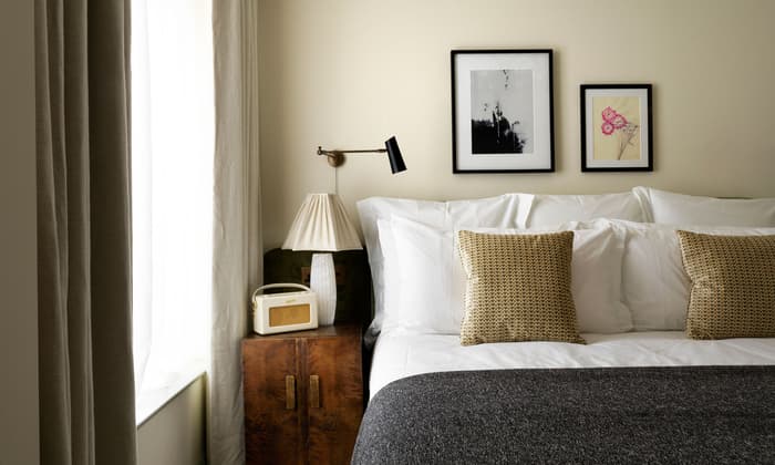 A neatly made bed with white linens and patterned pillows sits alongside a wooden nightstand holding a lamp and radio, adjacent to a large window with light curtains. Two framed artworks hang above the bed.