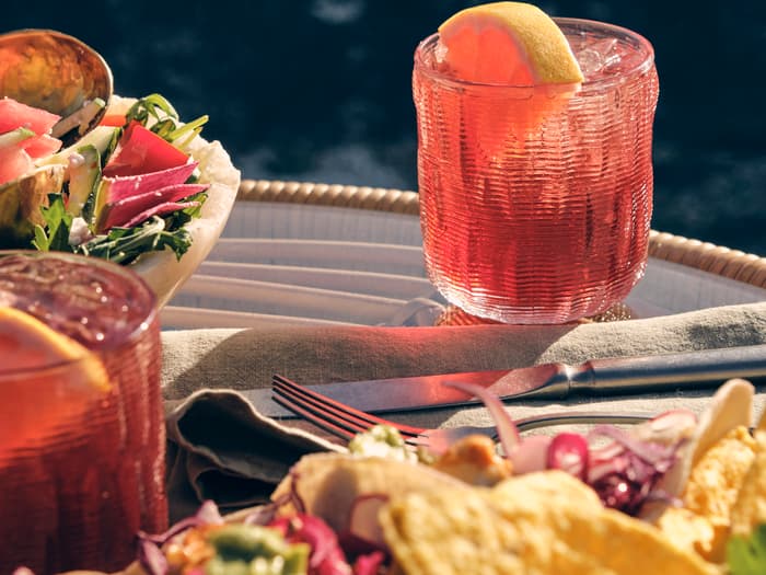 A red cocktail with an orange slice sits on a table next to a fork, knife, linen napkin, a salad, and tortilla chips, beside a dark, reflective water surface.