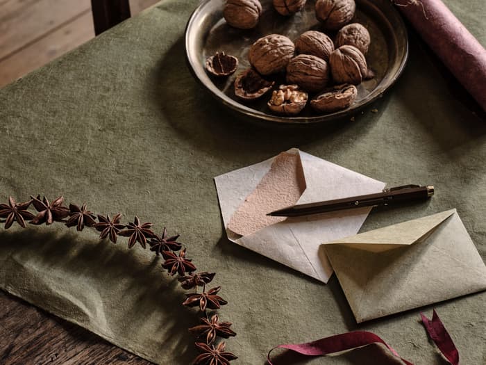 A wooden table holds uncracked walnuts in a metal bowl, scattered stationery including envelopes and paper, a pencil, a star anise wreath, and a purple ribbon, set in a rustic room.