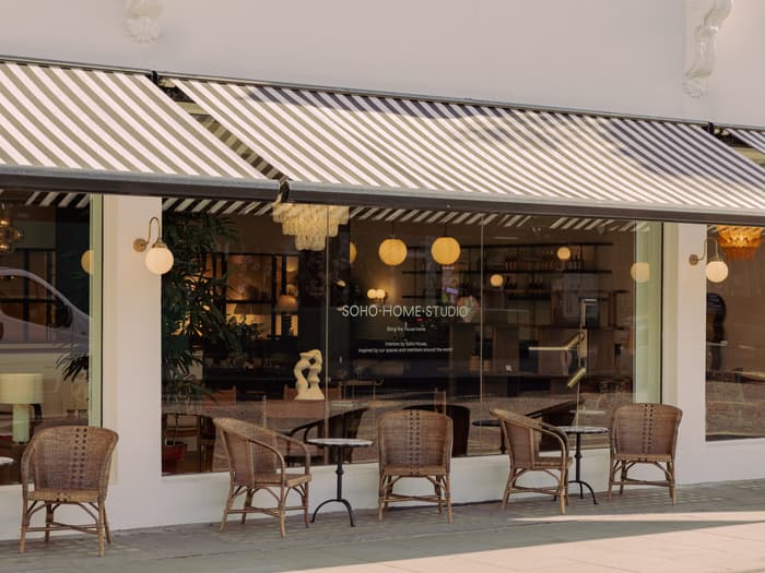 Wicker chairs and small tables sit outside a shop with striped awnings. The glass displays a sign reading: "SOHO HOME STUDIO. Bring the house home. inspired by Soho House." Inside, warm lighting and decor are visible.