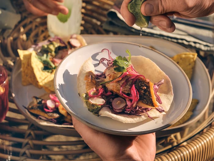 A hand squeezes a lime over a taco on a plate, while another hand holds a drink in a wicker outdoor setting with other food dishes around.
