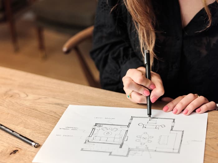 A woman draws on an architectural floor plan with a pen at a wooden table, surrounded by drafting tools, in a well-lit office with chairs in the background.