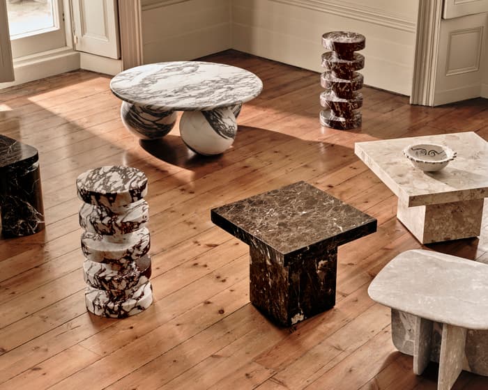 Marble tables of various shapes and sizes and stacked cylindrical sculptures rest on polished wooden floorboards in a brightly lit, elegant room with white doorways and large windows.