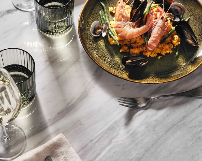 A plate of seafood paella with shrimp, clams, mussels, and green beans rests on a marble table next to three glasses of white wine, a fork, and a napkin.