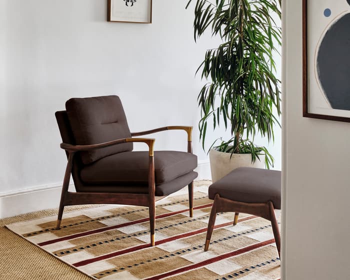 A dark brown armchair and matching footstool sit on a patterned rug in a white-walled room, adorned with two framed artworks and a tall potted plant.