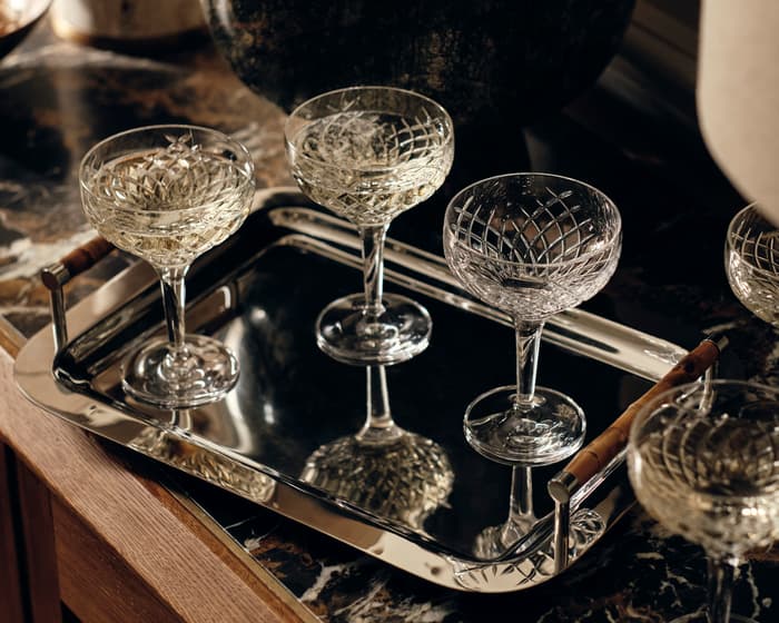 Five crystal champagne glasses rest on a reflective silver tray on a marble countertop, surrounded by a vase and a lamp in a dimly lit setting.