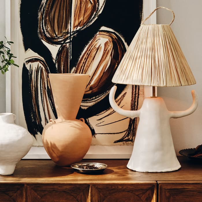 Three ceramic vases and a bowl sit on a wooden console table beside a leafy plant. A framed abstract painting with earthy tones hangs behind them, enhancing the stylish arrangement.